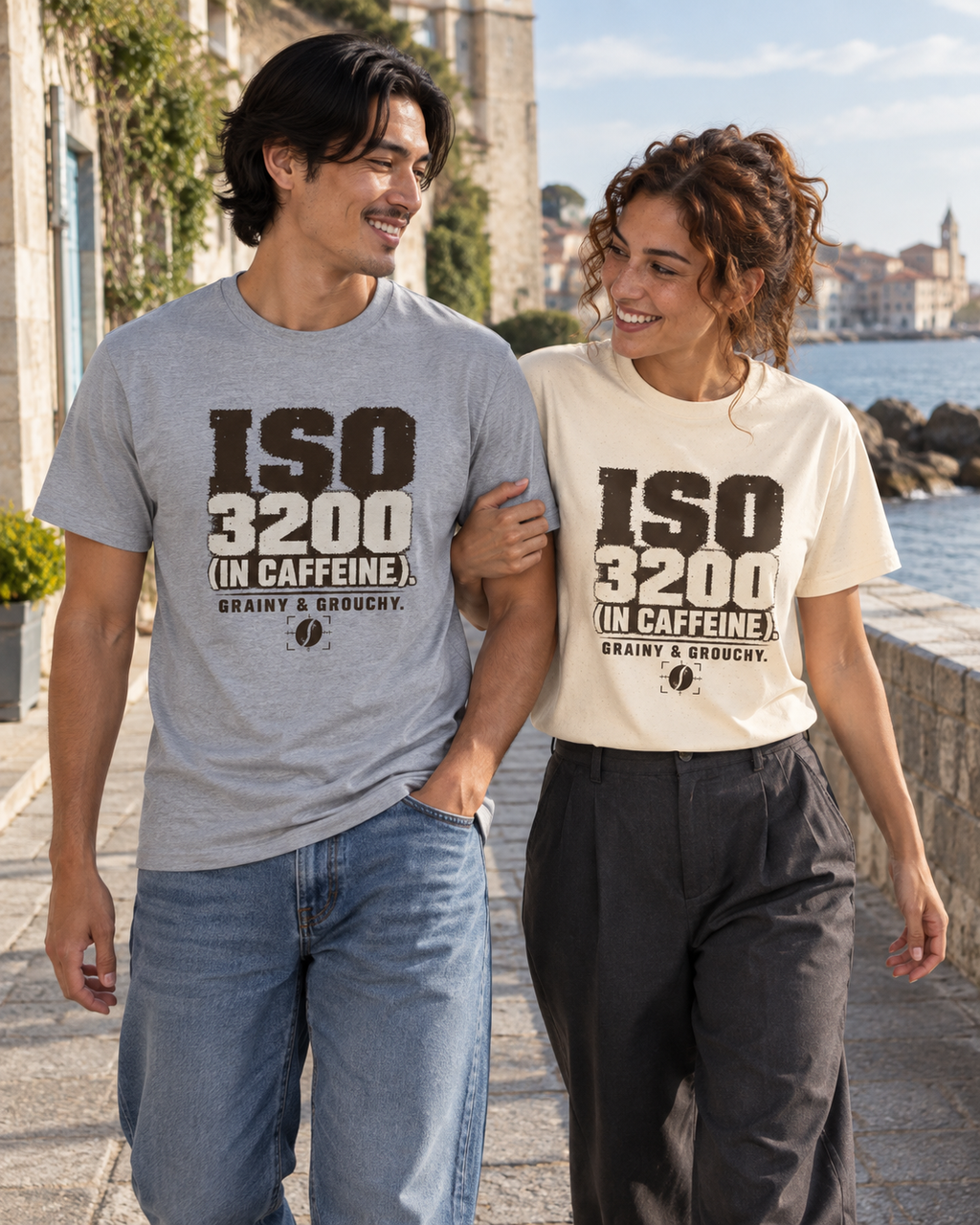 Two models walking arm-in-arm outdoors by the sea, wearing matching graphic t-shirts. The man wears a heather gray t-shirt with bold black text reading 'ISO 3200 (IN CAFFEINE) GRAINY & GROOVY', and the woman wears a cream-colored t-shirt with the same text.
