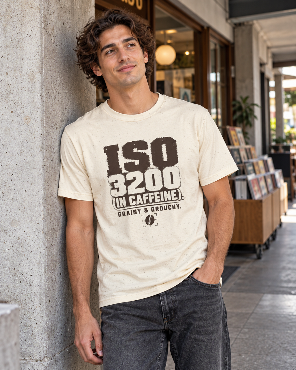 A man wearing a cream-colored graphic t-shirt with bold black text reading 'ISO 3200 (IN CAFFEINE) GRAINY & BROUGHERY.' leans against a concrete pillar in an outdoor cafe setting.