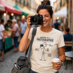 A woman in a street market holding a camera to her face, wearing a beige graphic T-shirt that reads 'ANATOMY OF A PHOTOGRAPHER' with a black line drawing. She has a gray shoulder bag and holds a coffee cup, with a blurred background of market stalls and people.