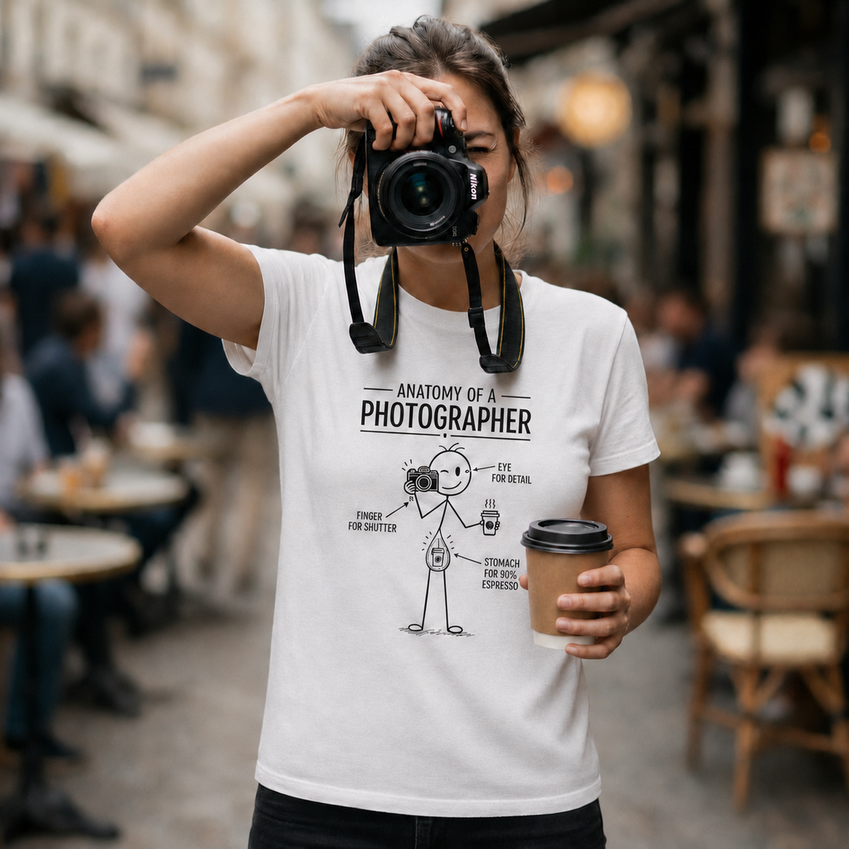 Woman photographer wearing an Anatomy of a Photographer T-shirt while holding a Nikon camera and takeaway coffee in a café.