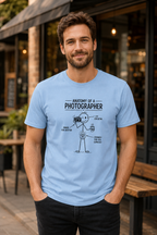 A man wearing a light blue graphic t-shirt with a black line art illustration titled 'ANATOMY OF A PHOTOGRAPHER' on the chest, standing outdoors in front of a cafe.